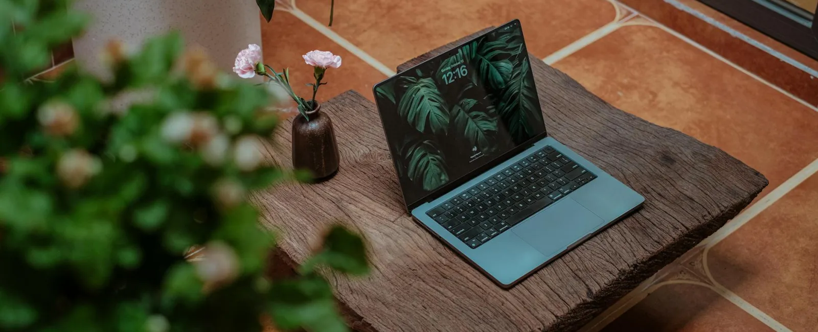 Modern laptop with green leaf wallpaper on wooden table next to flower vase and plant indoors