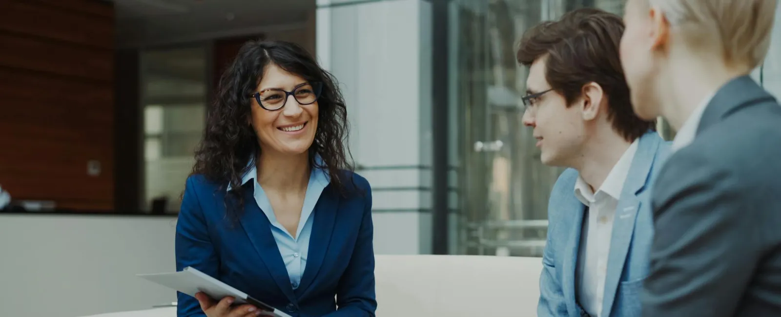 Smiling businesswoman holding documents while discussing with two male colleagues in modern office lounge.