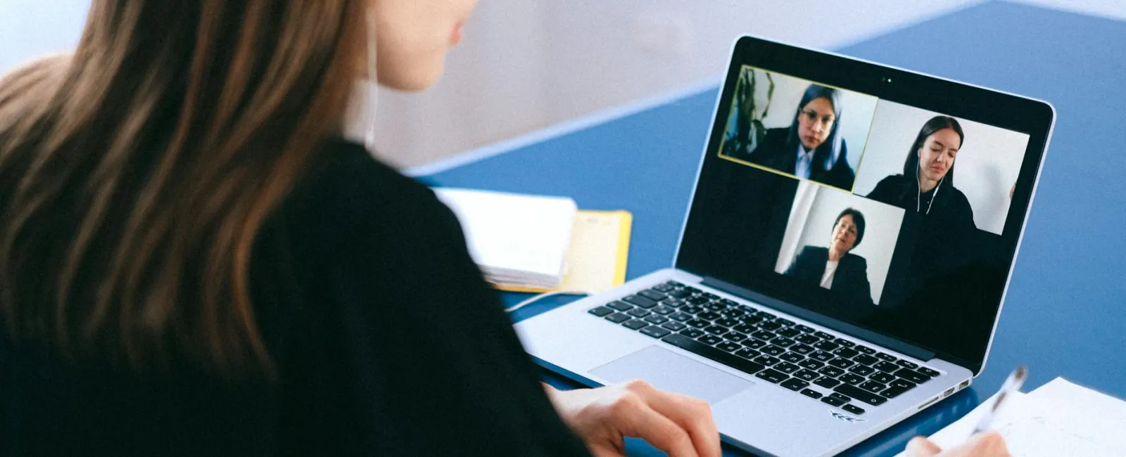 Woman attending virtual meeting on laptop while taking notes at blue desk in bright room