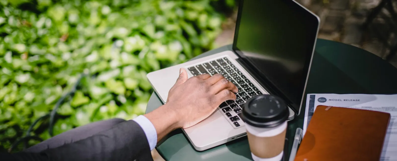 Person in suit working on laptop outdoors with coffee, notebook, AirPods, and documents on table.