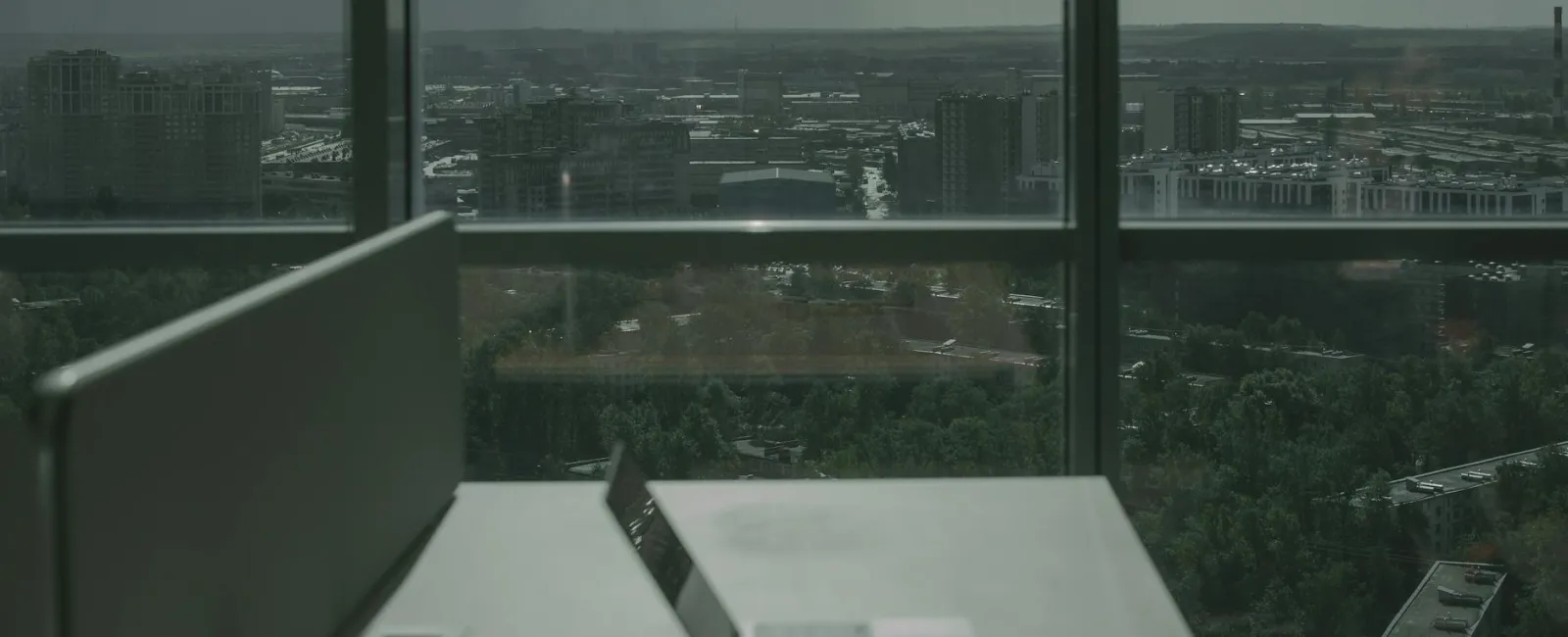Empty office desk with laptop overlooking a cityscape through large glass windows on a cloudy day