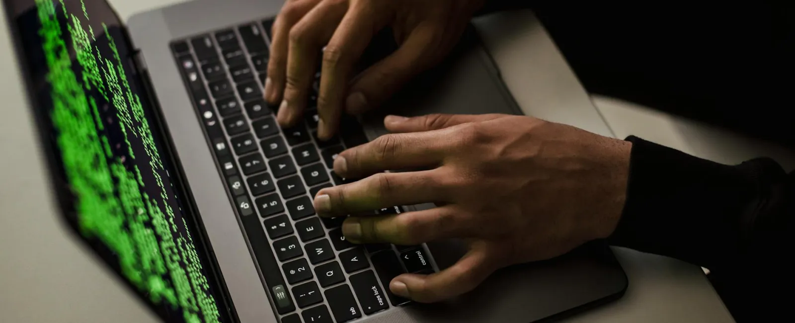 Hands typing on a laptop keyboard with green code on the screen and a wireless mouse nearby.