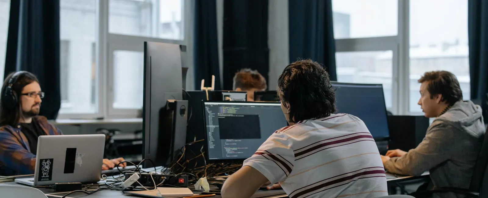 Four software developers working on computers in a modern office with large windows and black curtains