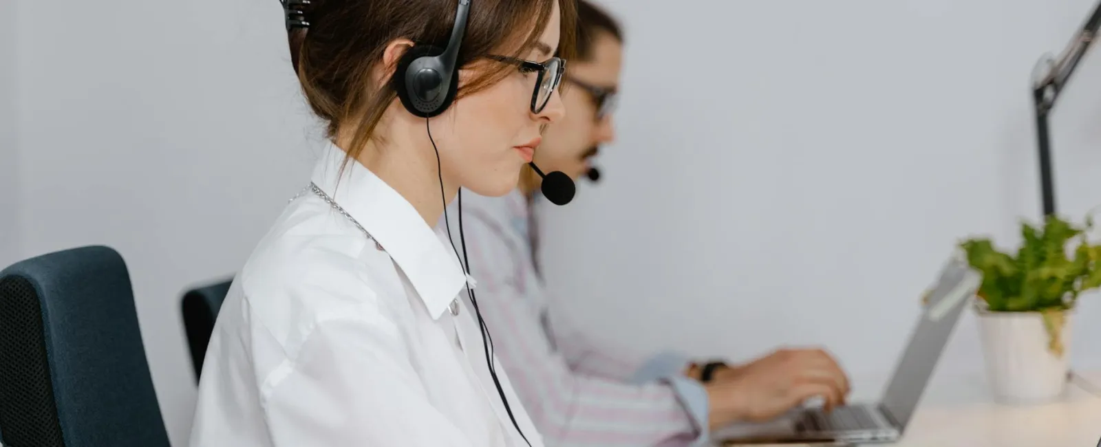 Two call center agents wearing headsets working on laptops in a modern office setting.
