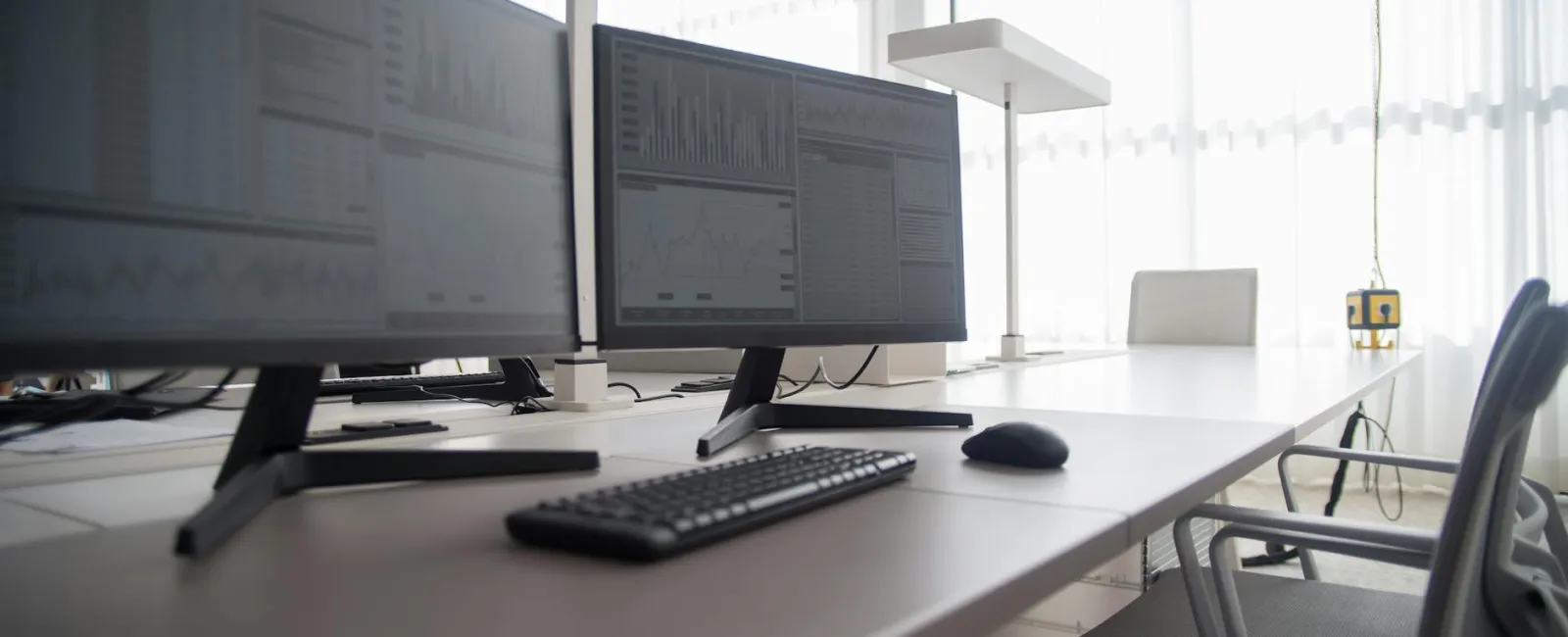Modern office desk with dual monitors displaying data charts, keyboard, mouse, and ergonomic chairs.