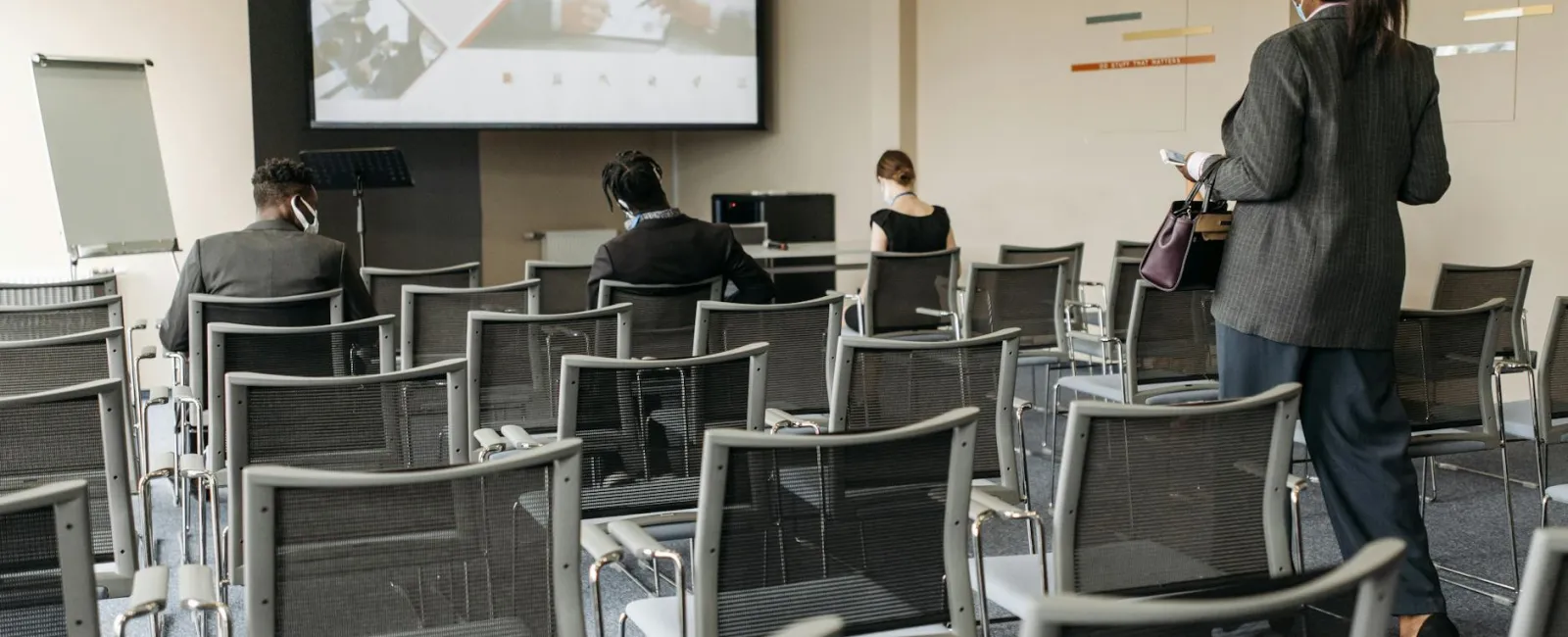 Business professionals wearing masks attend a socially distanced presentation on business analysis in a conference room.