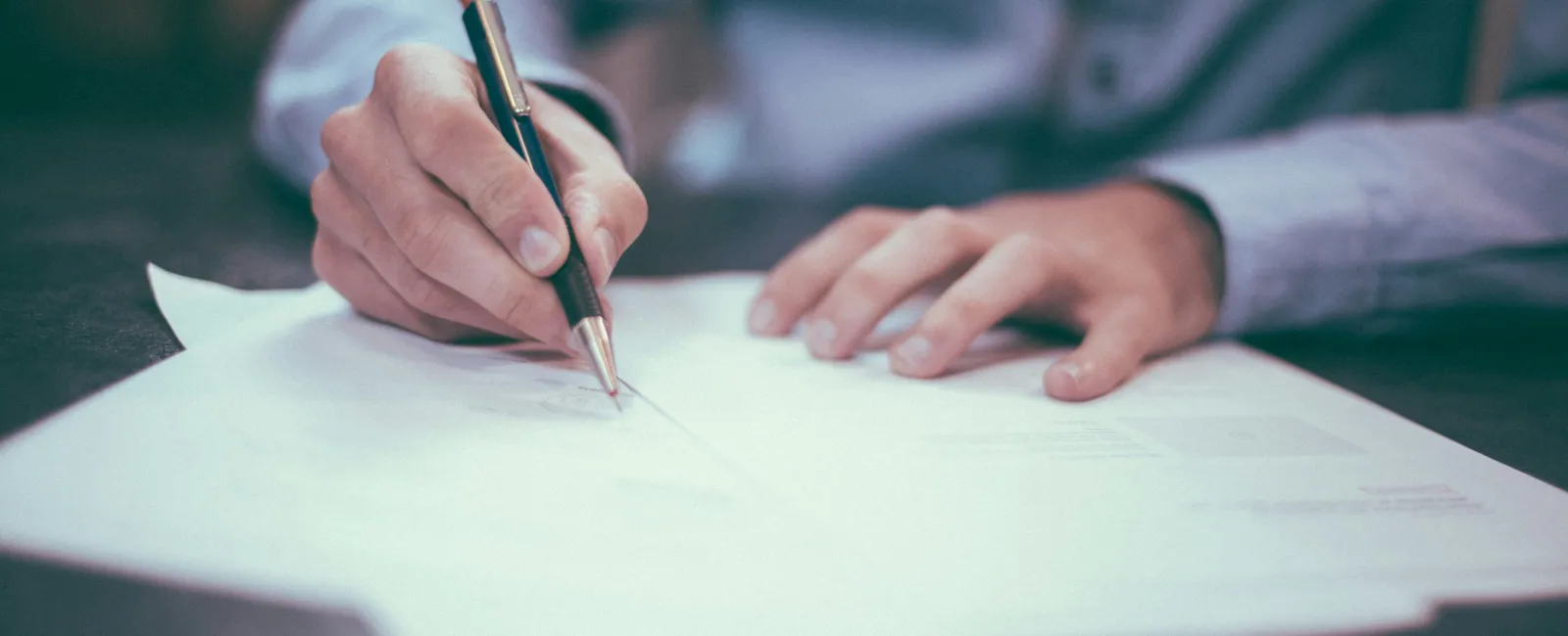 Person signing or writing on white paper documents with a pen on a desk in an office setting