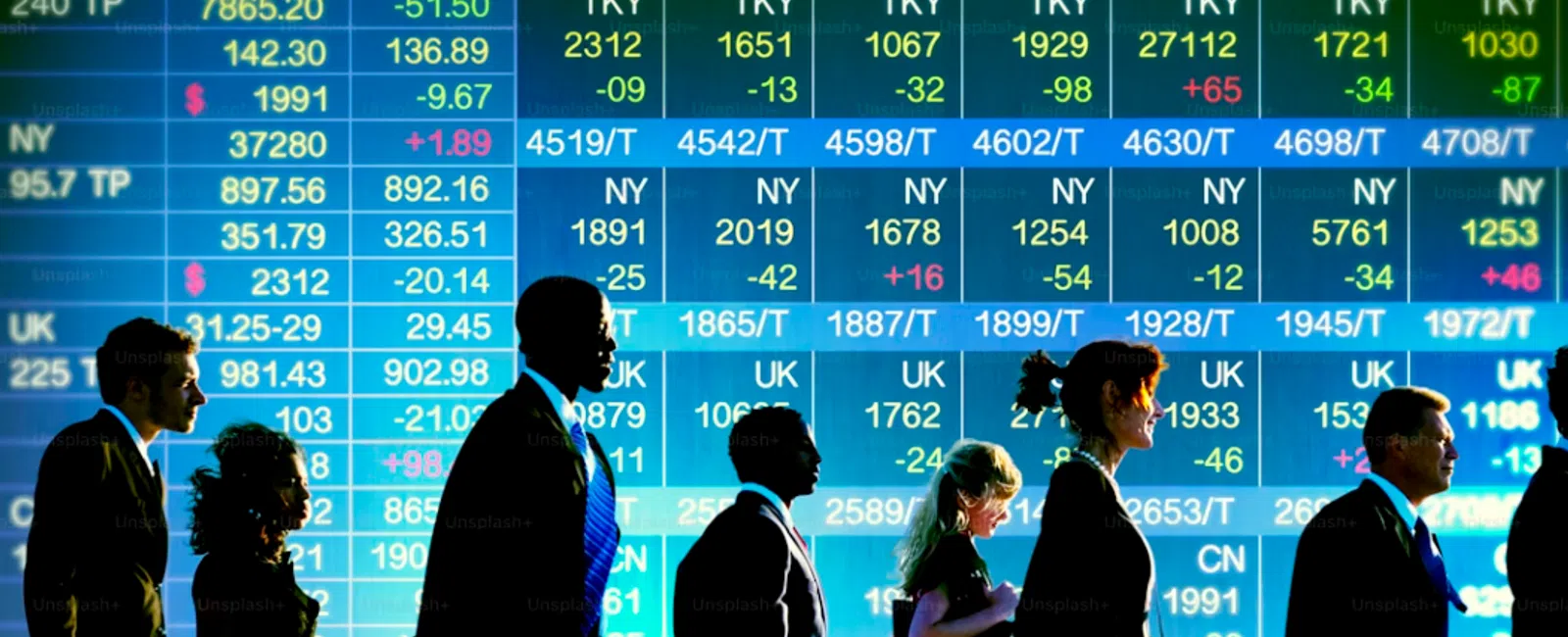 Silhouettes of business people walking in front of a large digital stock market data screen with numbers and symbols.
