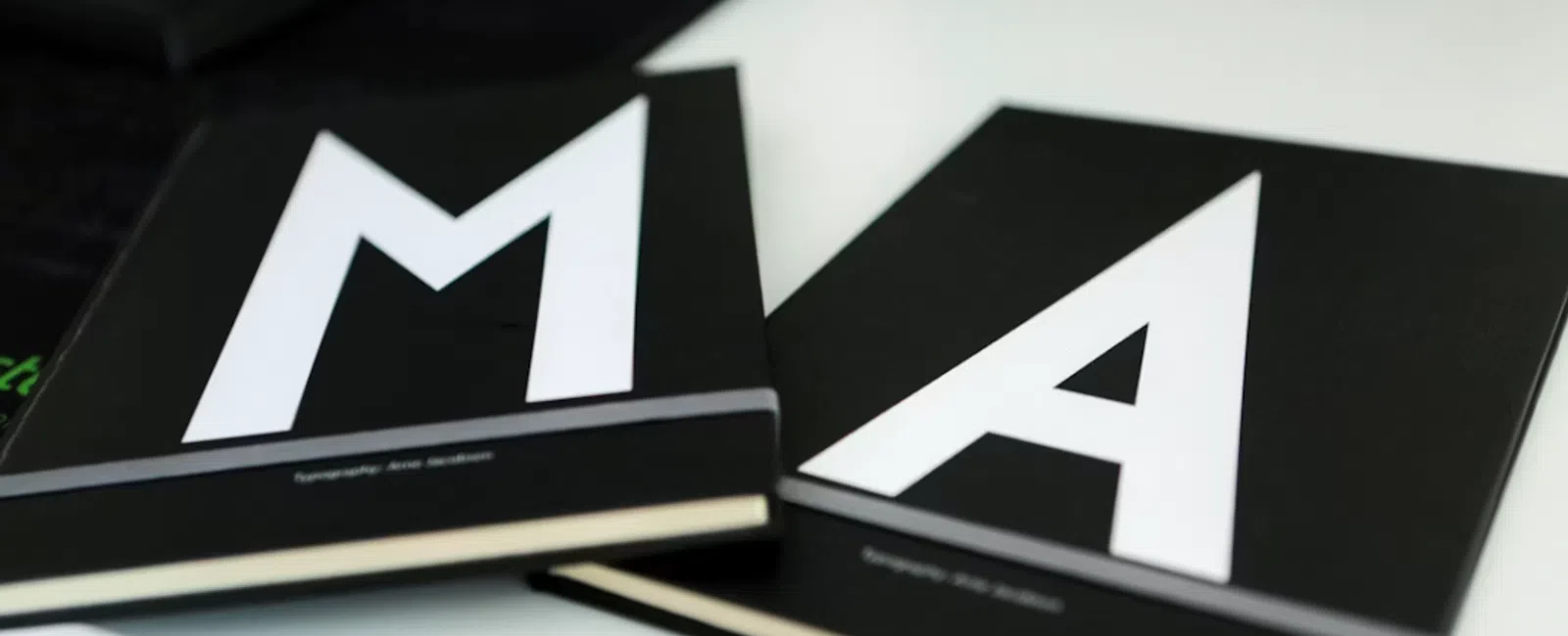 Black notebooks with large white letters M and A on the covers placed on a white surface.