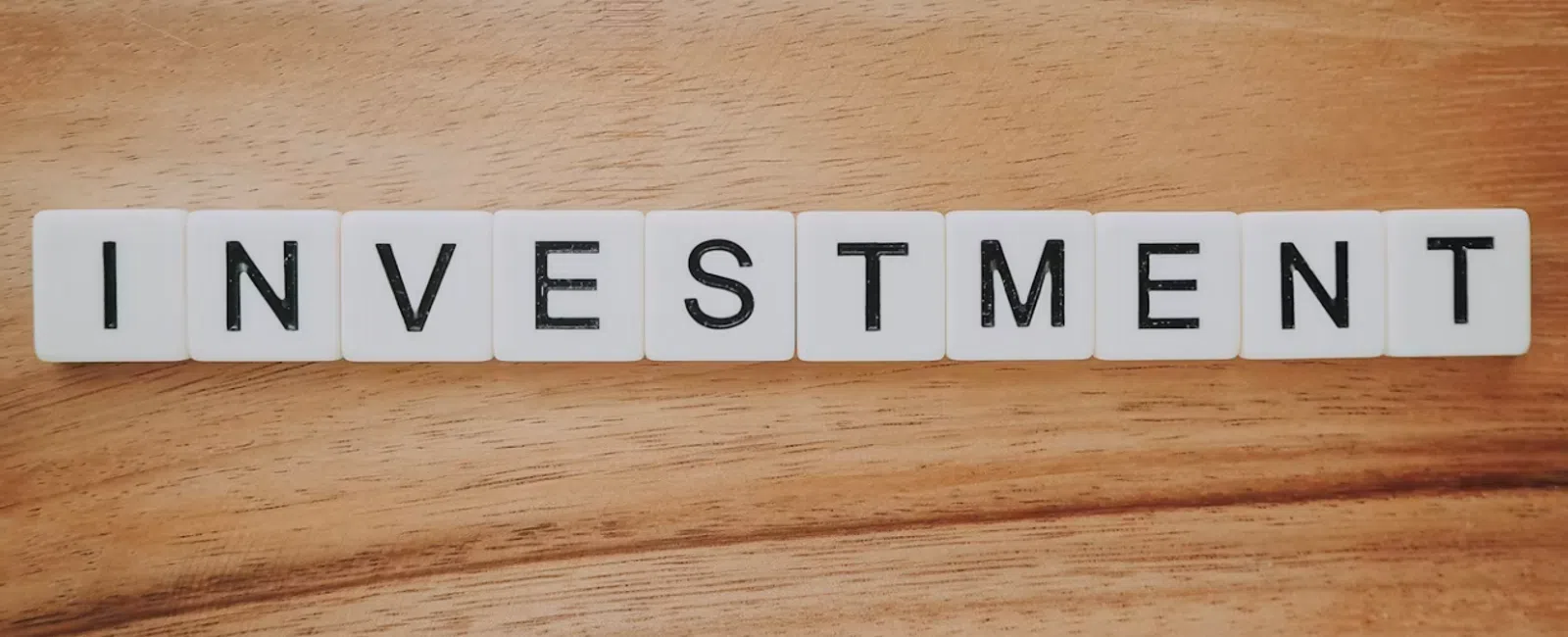 White tiles spelling the word INVESTMENT on a wooden surface indicating finance and money growth.