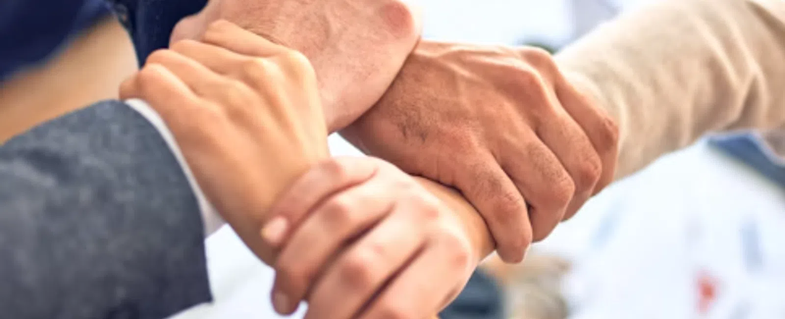 Four diverse hands holding each other's wrists in a teamwork gesture over a blurred office background