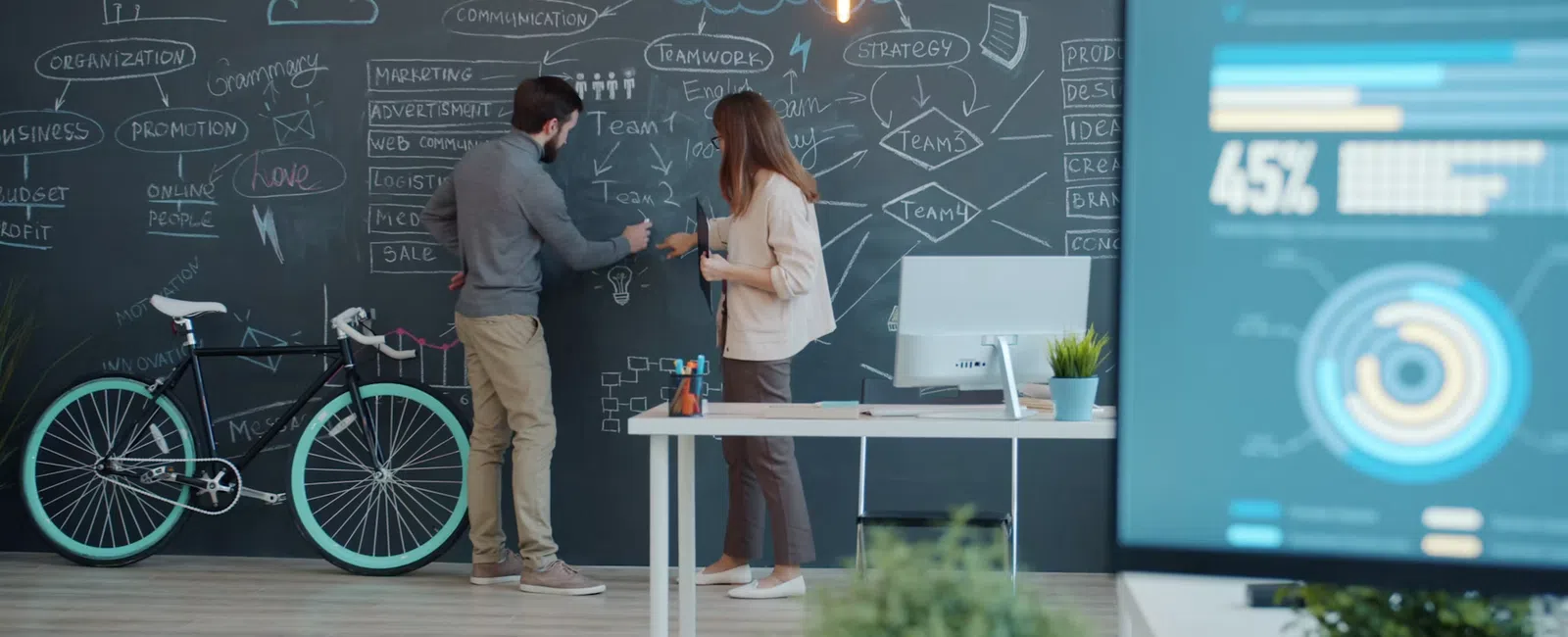 Two professionals brainstorming with charts and diagrams on a blackboard in a modern office workspace.