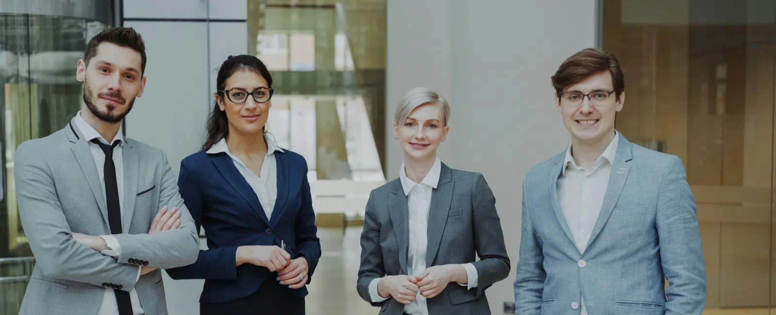 Four diverse business professionals in formal attire standing confidently in a modern office lobby.