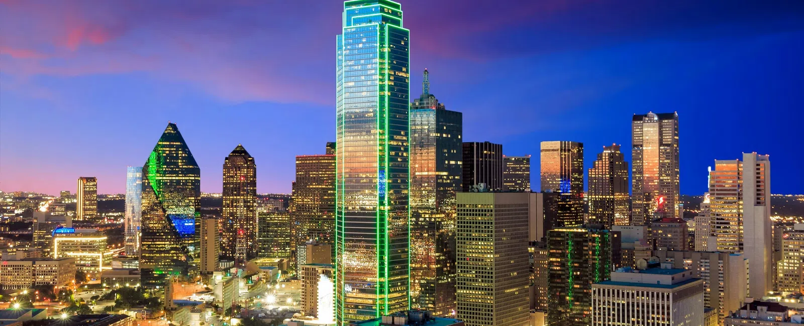 Dallas skyline at dusk with illuminated buildings, including the green-lit Bank of America Plaza tower.