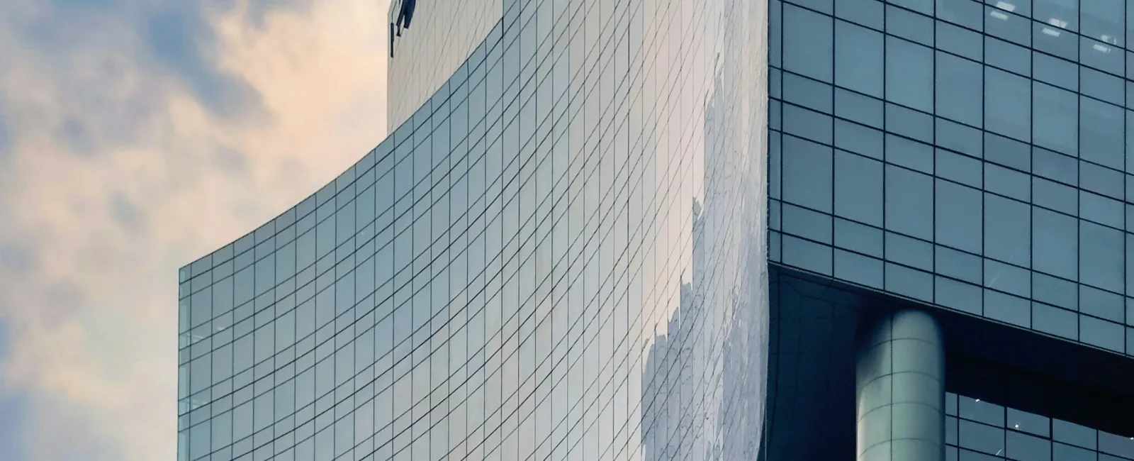 Curved modern glass skyscraper with reflections of sky and clouds against soft sunlight.