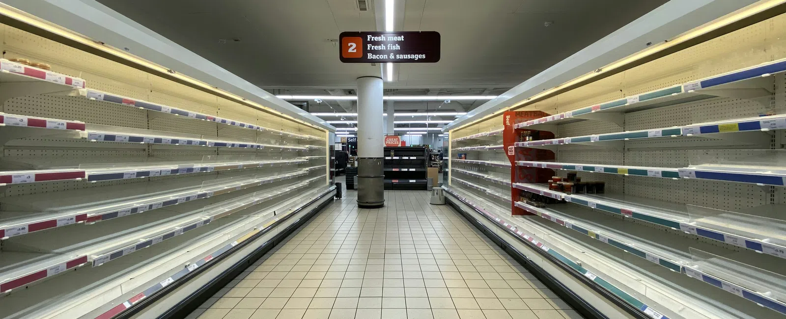 Empty refrigerated shelves in a supermarket aisle labeled for fresh meat, fish, bacon, and sausages.