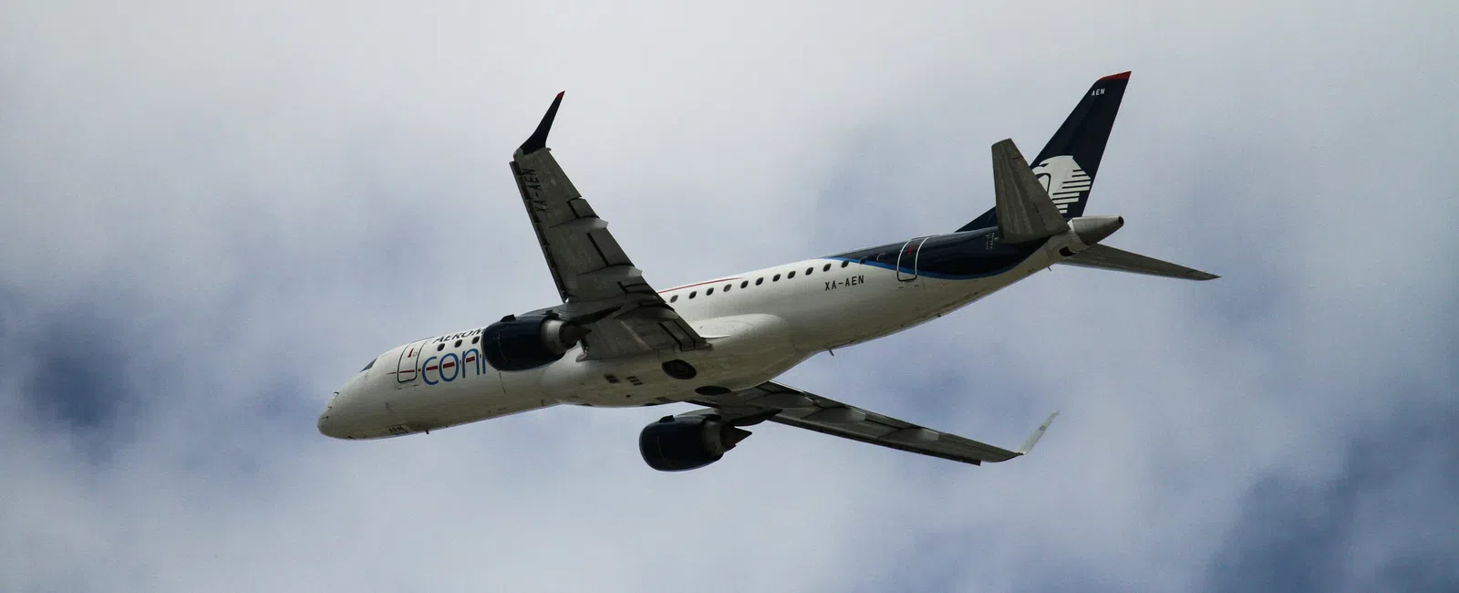 Commercial airplane flying in cloudy sky with blue and white patterns visible under wings.