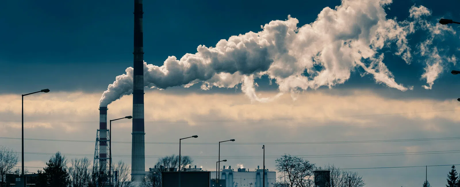 Industrial smokestacks emitting thick smoke over a city road with cars under a cloudy sky.