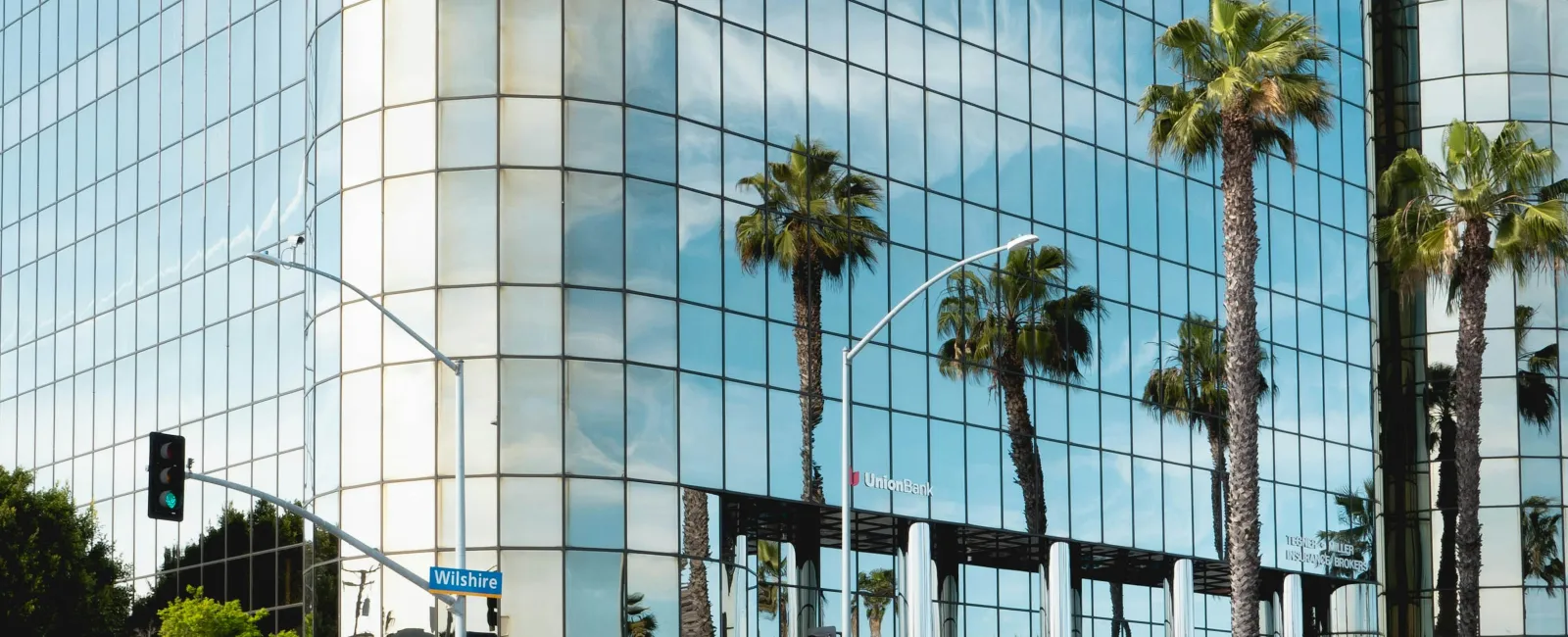 Modern glass office building reflecting palm trees and blue sky at Wilshire intersection with blurred car motion.