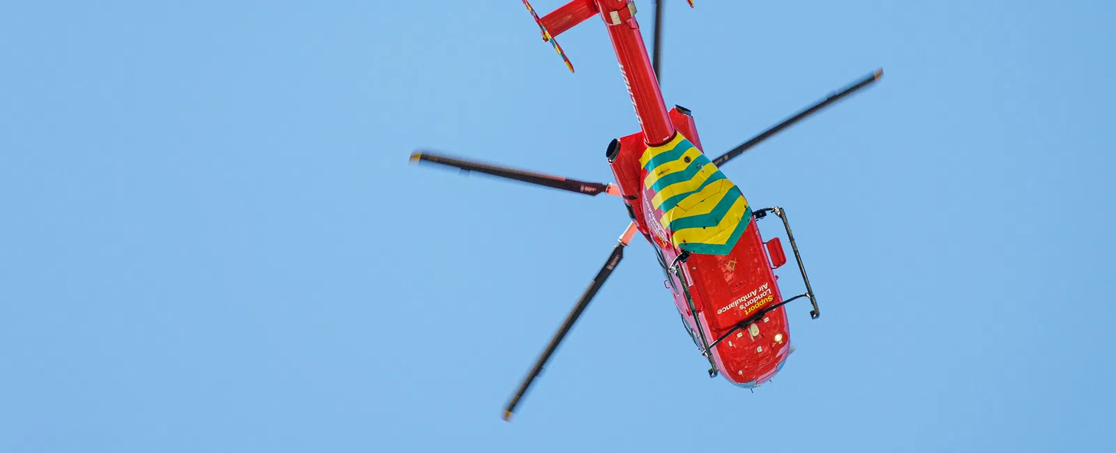 Red rescue helicopter with yellow and green markings flying against a clear blue sky.