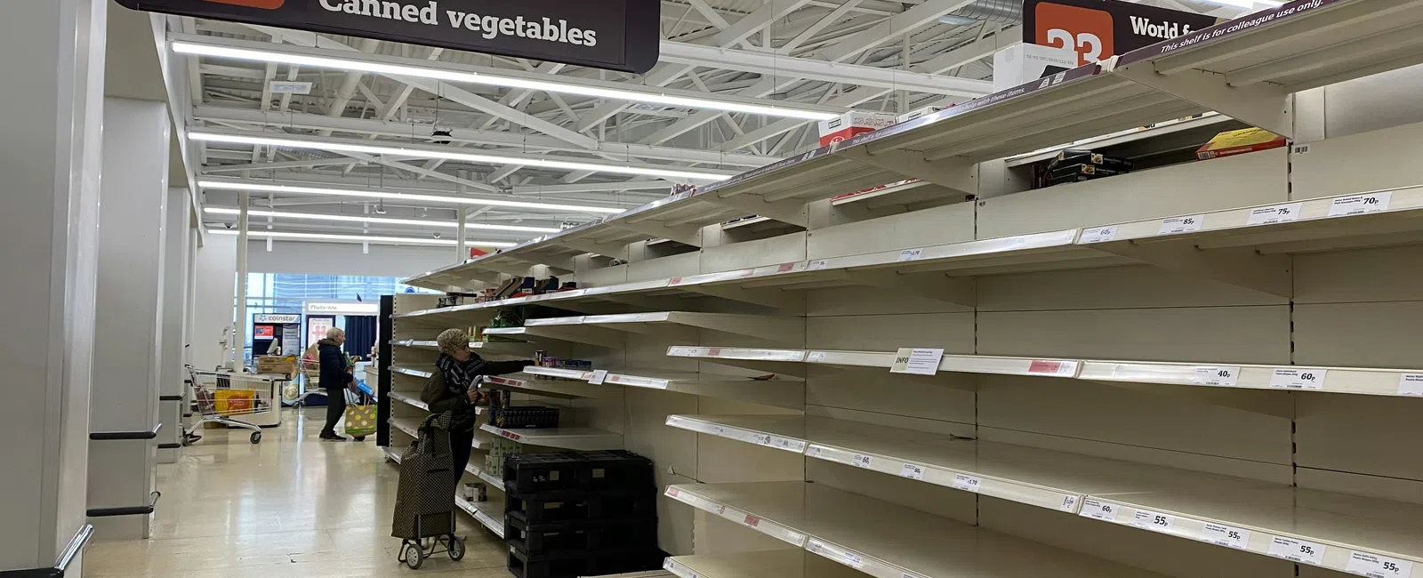 Empty grocery store shelves in aisle 22 for canned soup, beans, and vegetables with few shoppers.