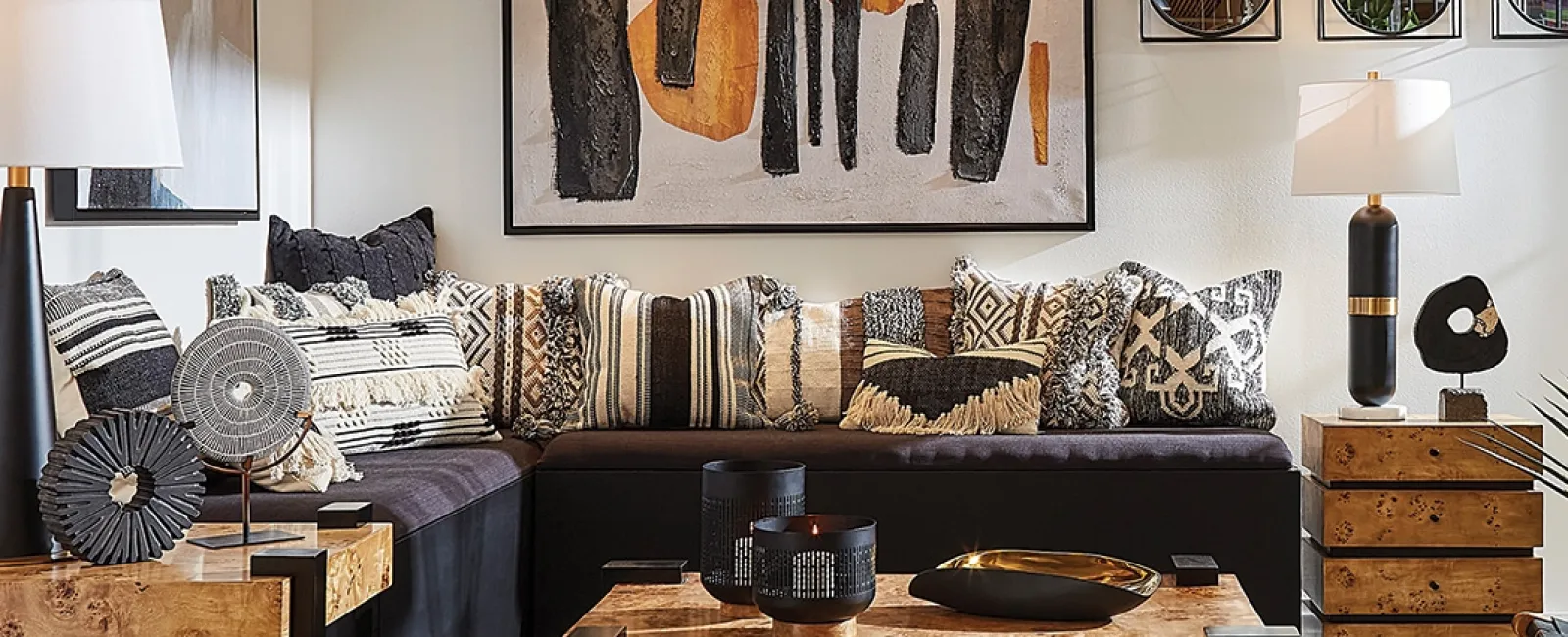 Modern living room with black sectional sofa, patterned pillows, wooden tables, abstract artwork, and contemporary lighting.