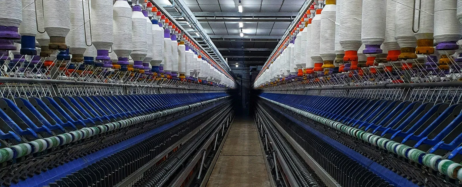 Long aisle between rows of textile spinning machines with white thread cones in a factory setting.