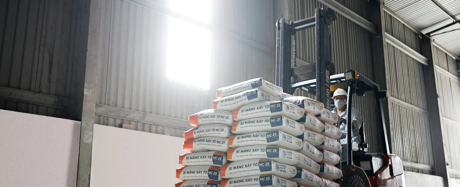 Forklift carrying stacked cement bags inside a warehouse with high windows and industrial walls.