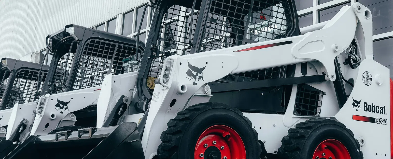 Row of white Bobcat S530 skid-steer loaders with red wheels parked outside a building under overcast sky