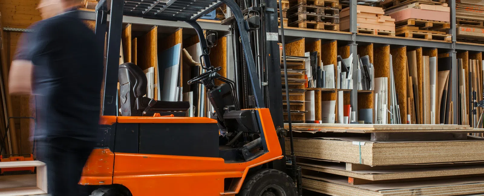 Orange forklift in wood warehouse with stacked plywood and pallets, blurred worker walking by.