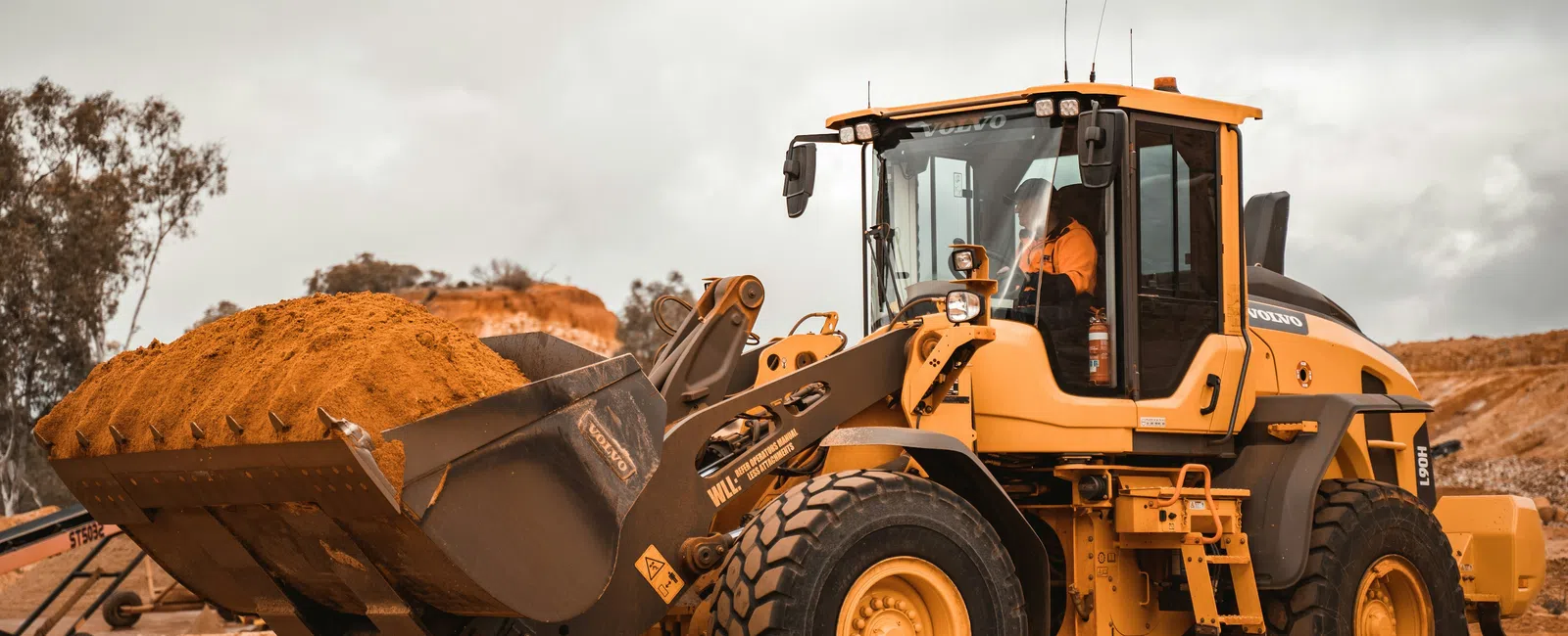 Yellow construction loader carrying dirt at a dusty excavation site under cloudy sky.