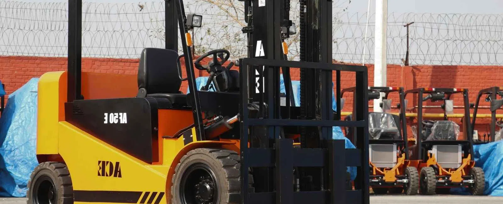 Yellow and black forklift parked outdoors with multiple forklifts visible in the background under clear skies.