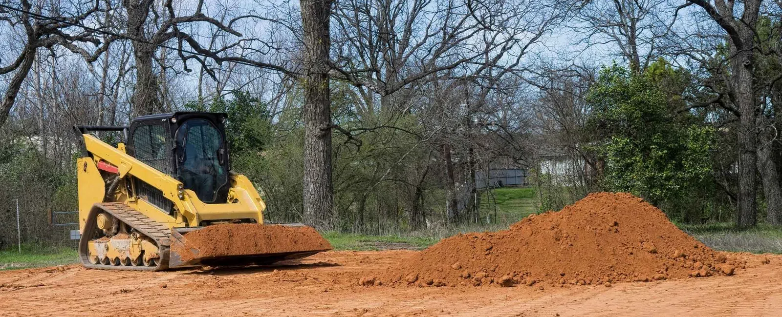 Yellow bulldozer leveling dirt on a cleared land with trees and blue sky in the background