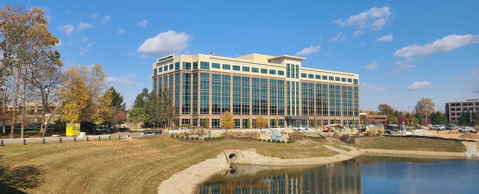 Modern office building with glass windows by a pond under a blue sky with autumn trees around.