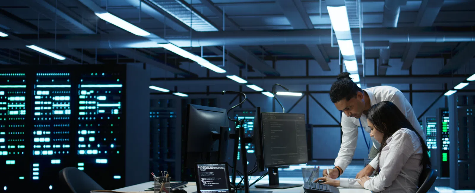 Two IT professionals collaborate on coding in a modern data center with servers and multiple monitors.