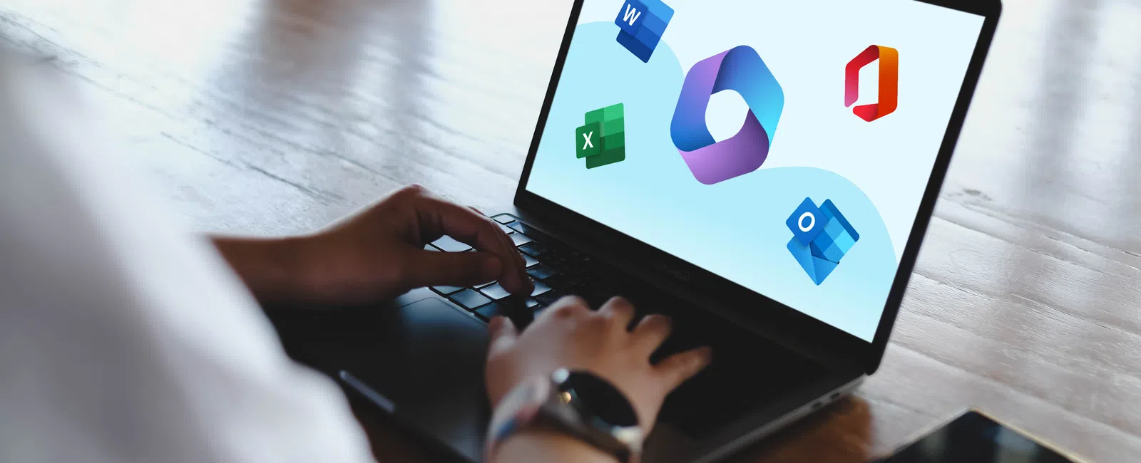Person using laptop displaying Microsoft Office apps icons on screen at wooden table near smartphone.