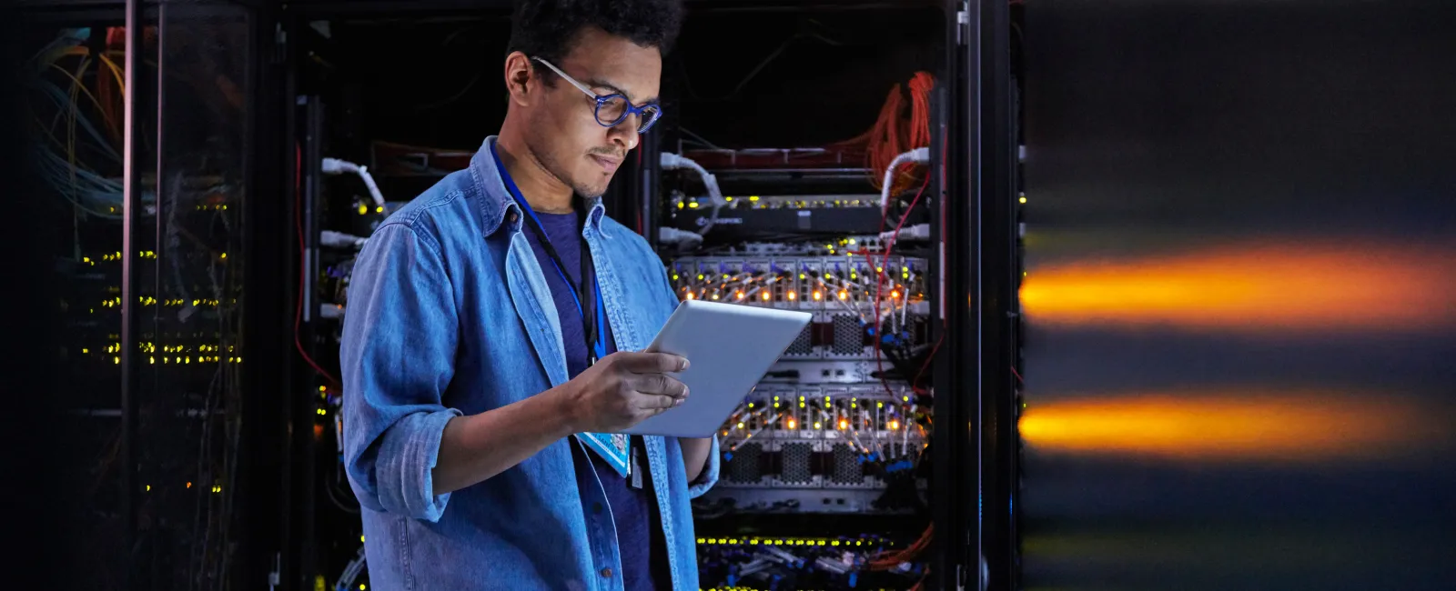 Technician in blue shirt using tablet to check server racks with glowing lights in data center room.