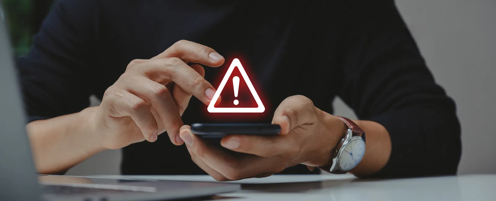 Person holding smartphone with digital red warning alert symbol above screen, laptop nearby on table.