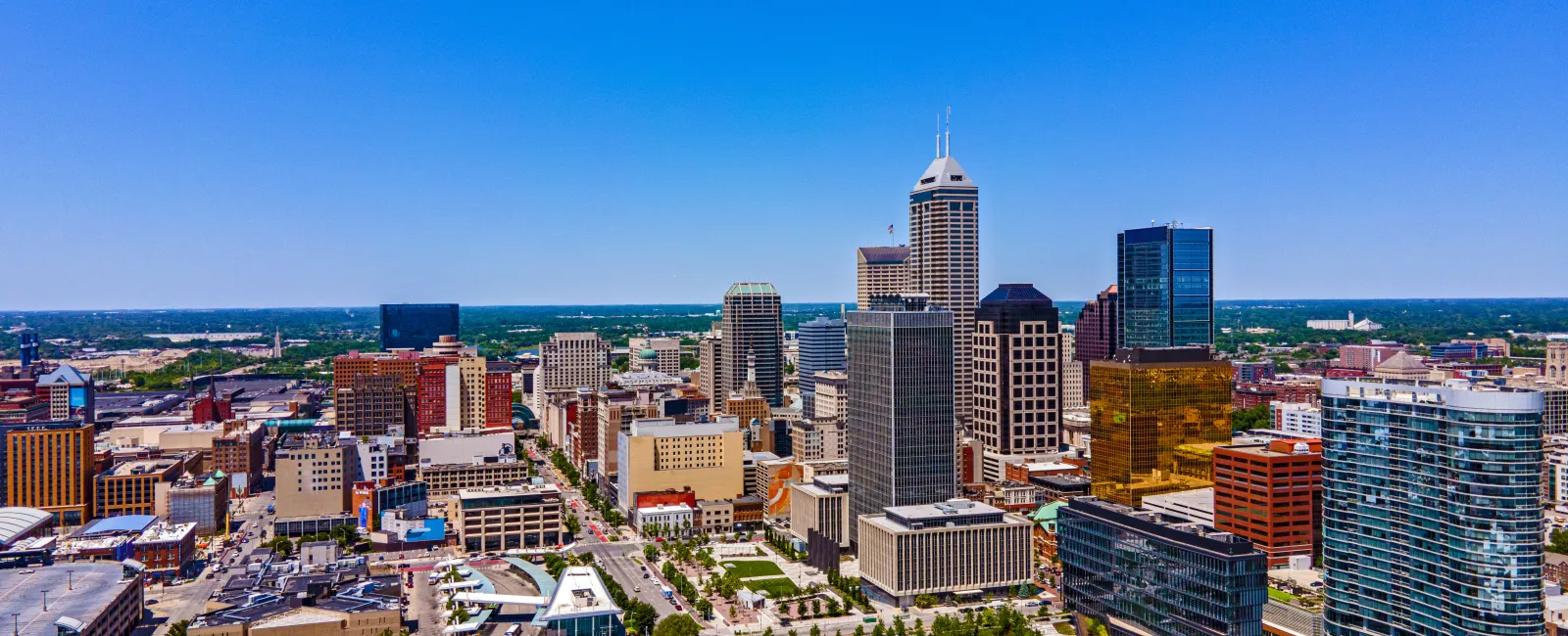 Aerial view of downtown Indianapolis featuring skyscrapers, green parks, and clear blue sky on a sunny day.