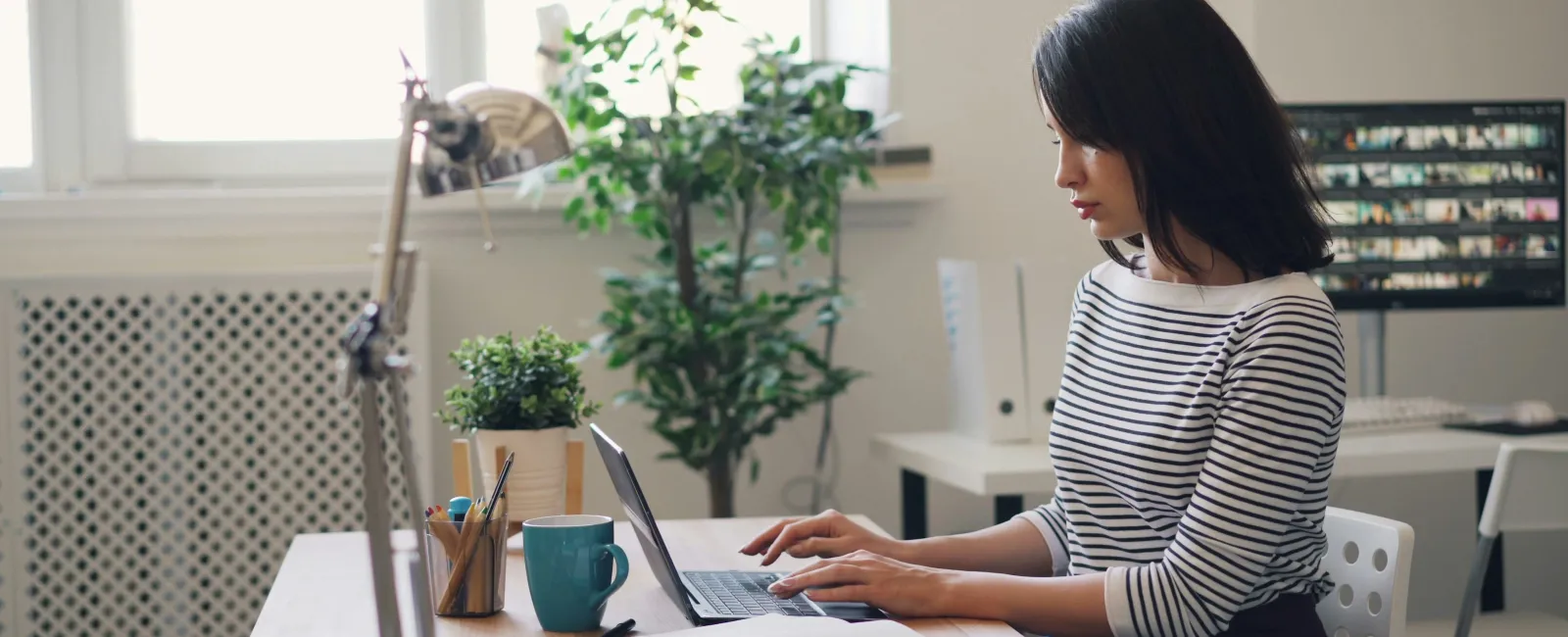 Young woman in striped shirt working on laptop at desk with plant, notebook, and coffee in bright office.