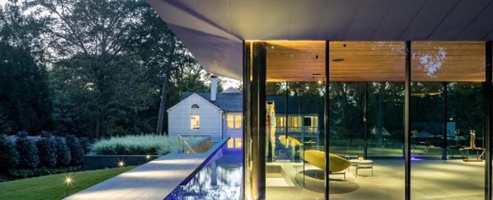 Modern glass house with pool lit by purple lights at dusk beside traditional white wooden home surrounded by trees.