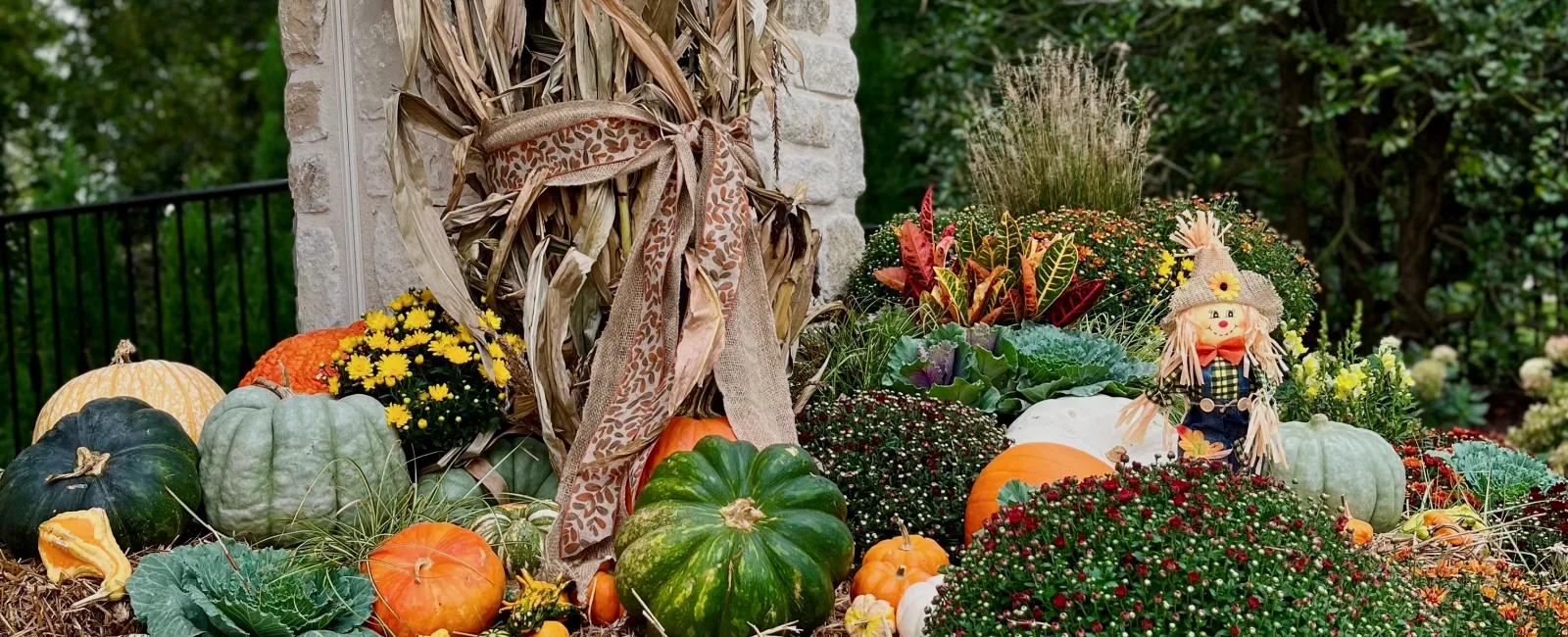 Colorful autumn display of pumpkins, gourds, mums, hay bales, and a scarecrow at a stone porch corner.
