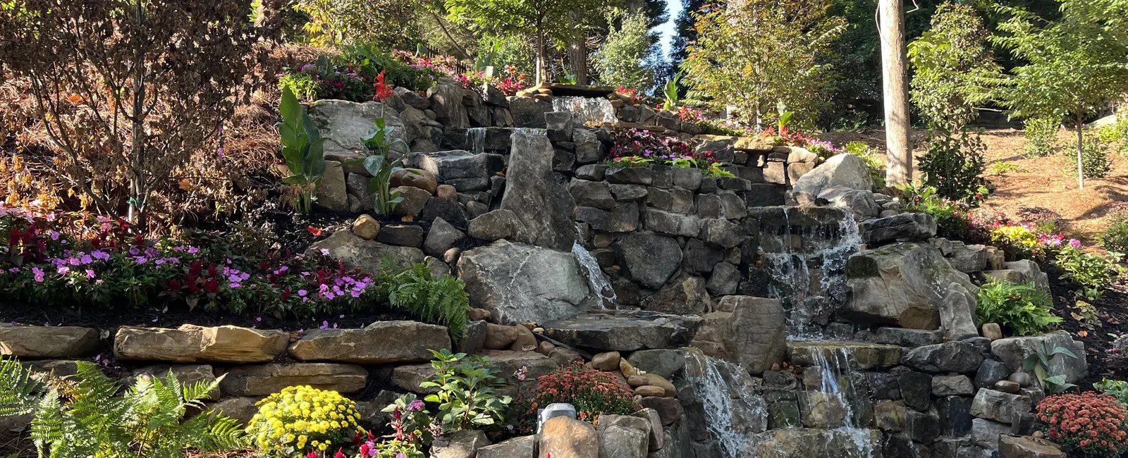 Multi-tiered rock waterfall surrounded by colorful flowers and lush greenery in a landscaped garden.