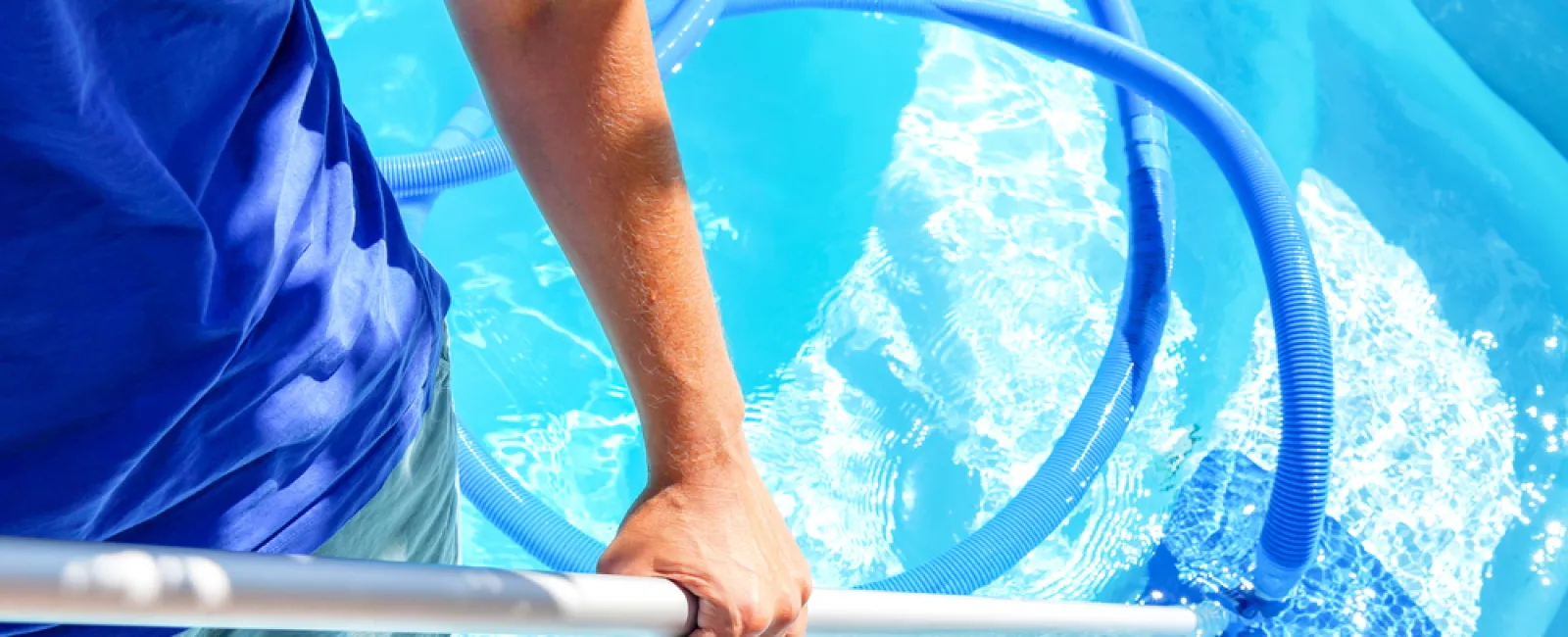 Person cleaning clear blue swimming pool water with a manual pool vacuum and hose on a sunny day
