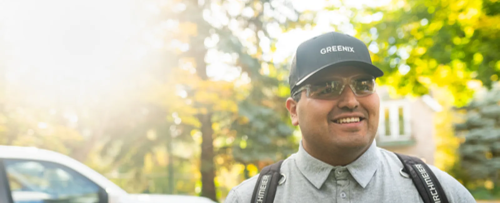 Smiling pest control technician with Greenix cap and uniform standing outdoors near a Greenix truck.