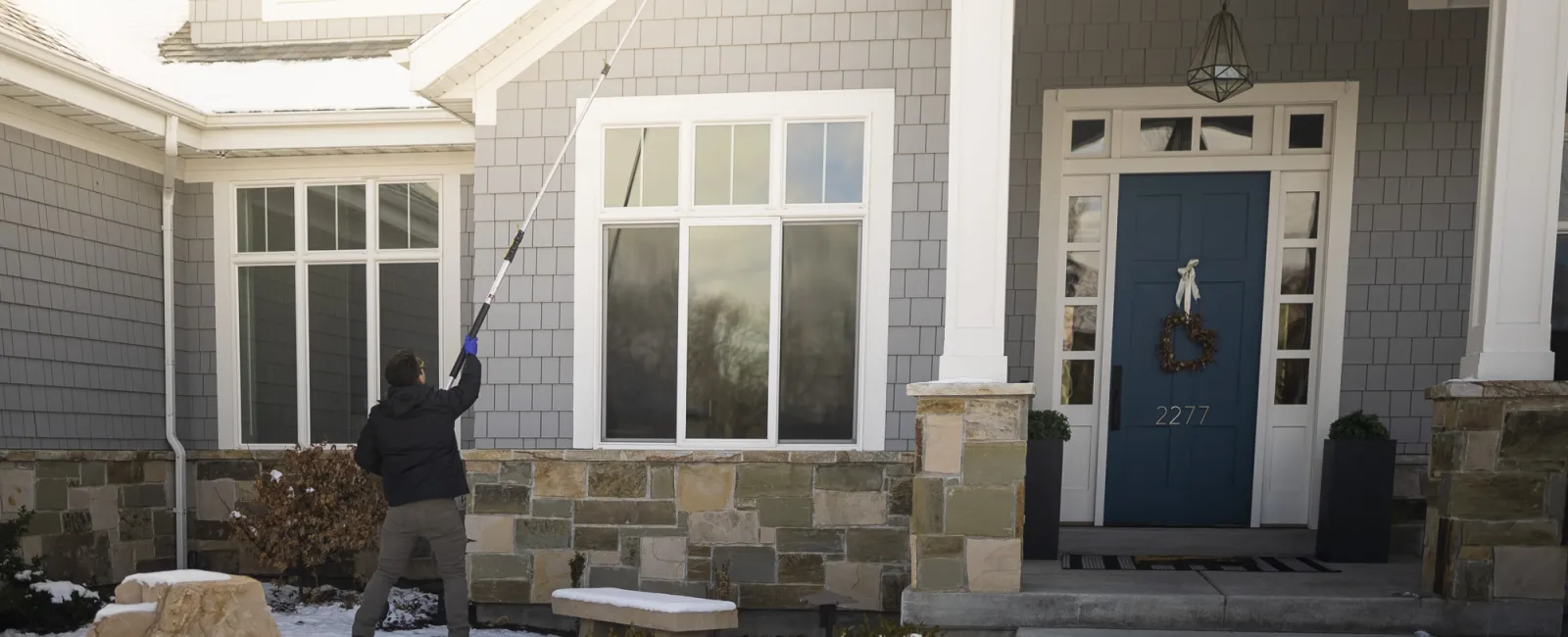 Person using a long tool to clean windows of a gray house with snow on the ground