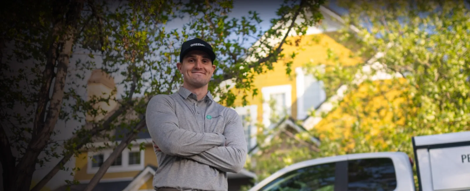 Confident technician with crossed arms standing outdoors near a white service truck and yellow house in sunlight.