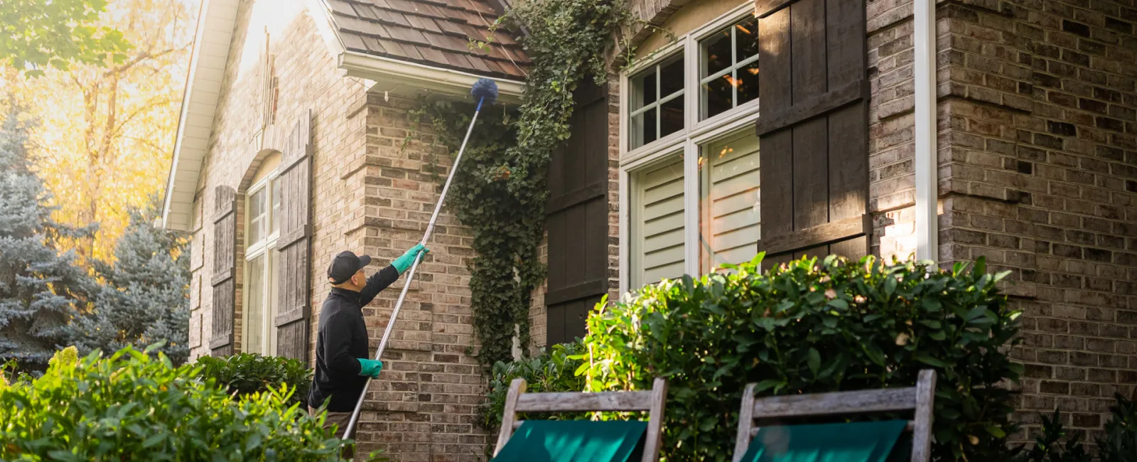 Person cleaning house exterior brick walls with a long brush during daylight surrounded by green plants and chairs.