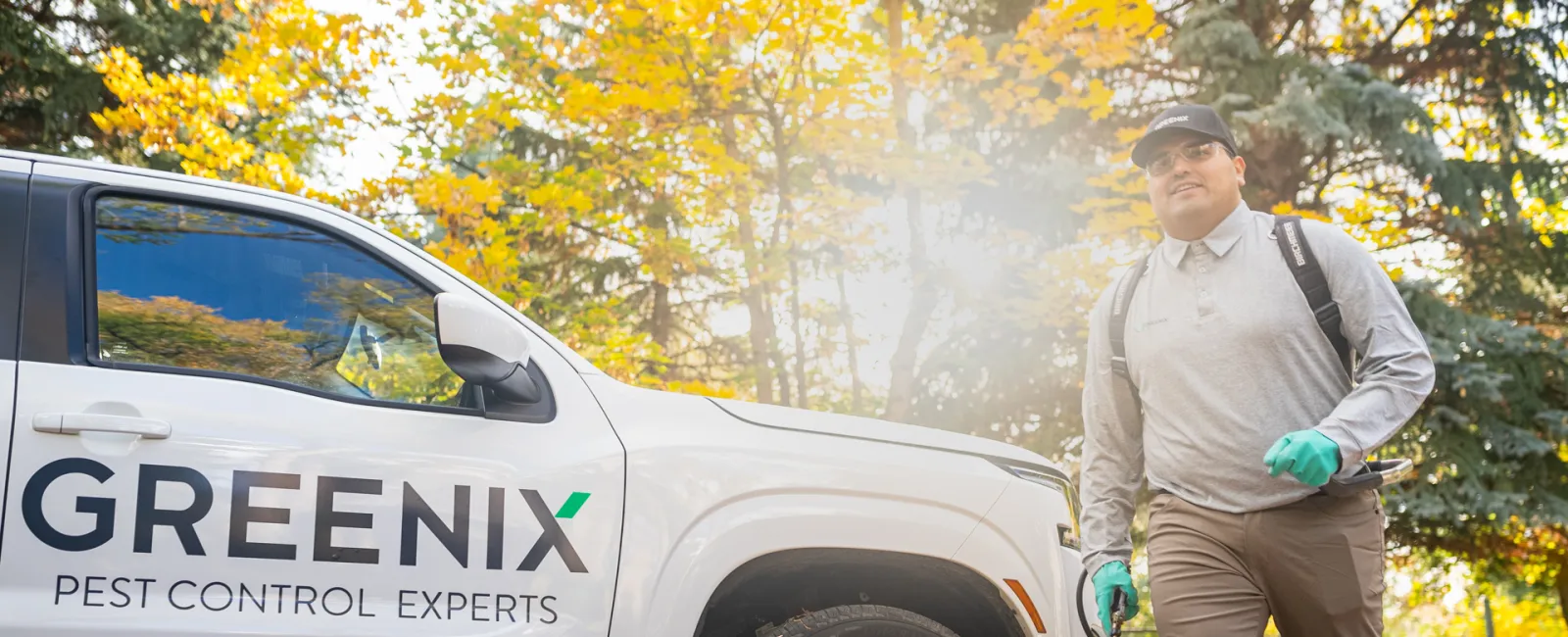 Pest control technician walking with equipment near a Greenix Pest Control Experts vehicle in autumn setting.