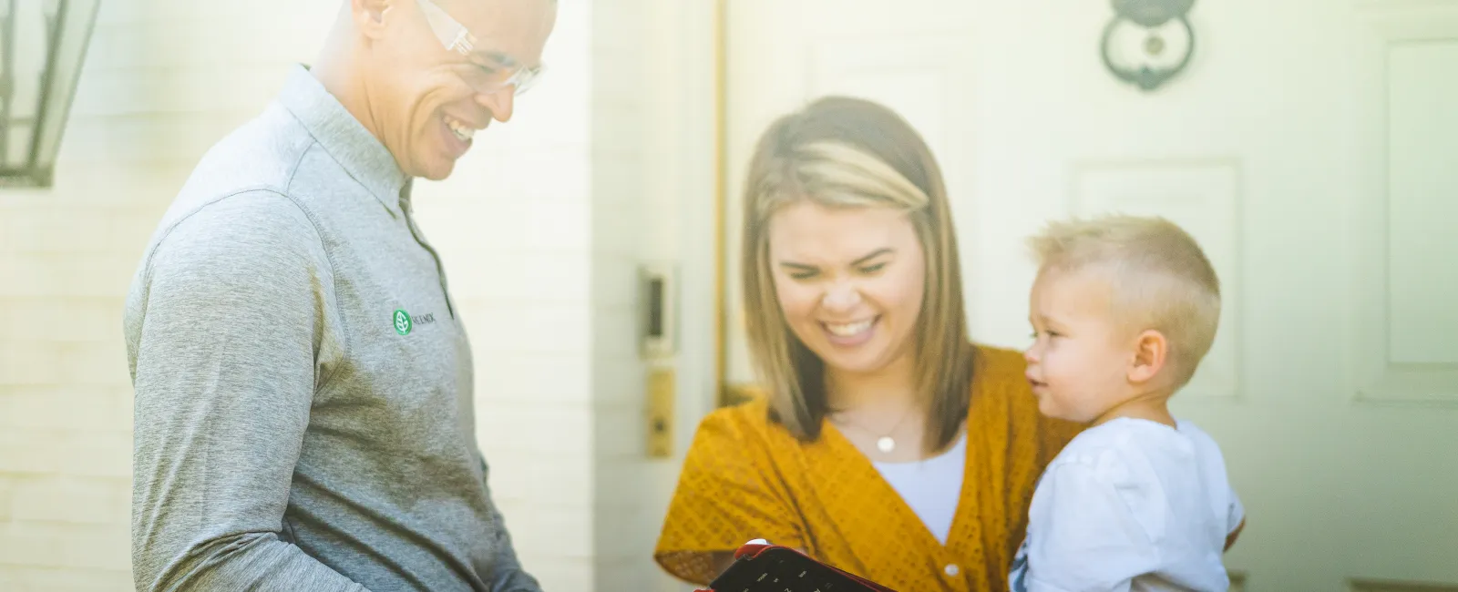 Technician showing tablet to smiling woman holding child outside a home during a service visit.
